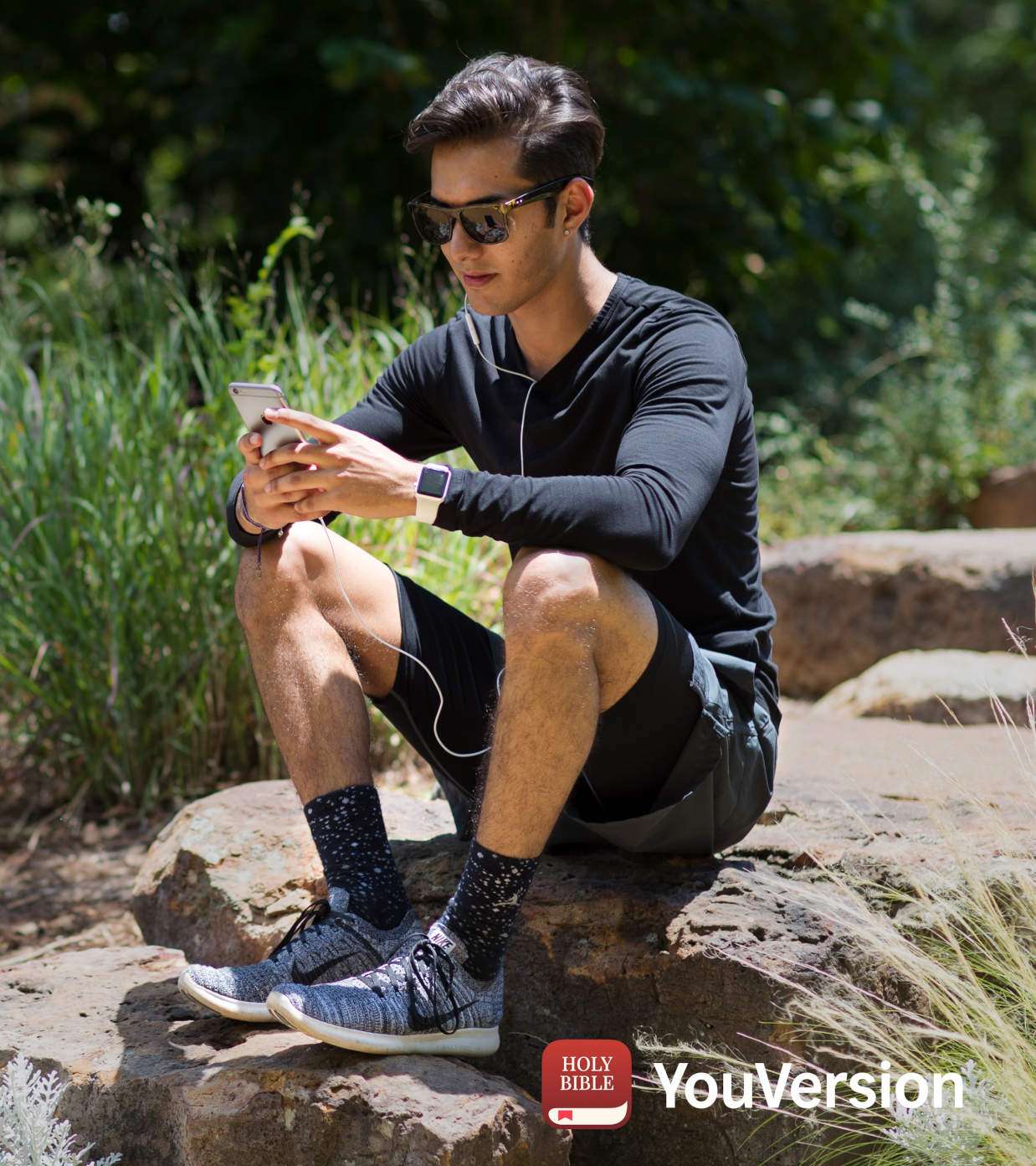 A man in sportswear sits on a boulder, holding a smartphone in his hand.